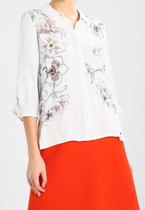 Weiße Bluse mit Knopfleiste und rosa-schwarzem Blumenmuster, getragen zu einem knallorangen Rock, zeigt oberen Torso und linken Arm.