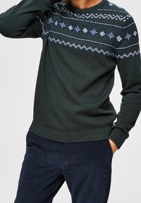 Mörkgrön stickad tröja med rund halsringning, prydd med vita och blå geometriska mönster över bröstet samt ribbade muddar och nederkant.