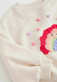 Cremefarbener Sweatshirt mit einem gestickten Löwenmotiv, verziert mit bunten Applikationen und Pom-Pom-Akzenten in Pink, Lila und Rot.