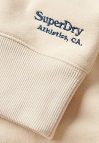 Tejido de sudadera beige con puños acanalados. Bordado en azul oscuro "SuperDry Athletics, CA" en el puño. Textura suave.