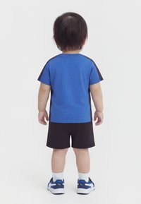 Παιδάκι που στέκεται γυρισμένο πλάτη, φορά μπλε και μαύρο κοντομάνικο μπλουζάκι, μαύρο σορτς, λευκές κάλτσες και μπλε και λευκά αθλητικά παπούτσια.