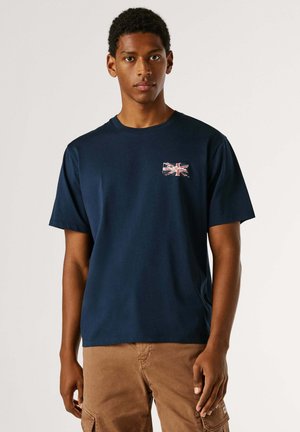 Giovane uomo che indossa una maglietta blu navy con una piccola bandiera britannica sul petto e pantaloni cargo marroni, in piedi contro uno sfondo semplice.