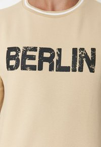 Camiseta beige con el texto desgastado en negro "BERLÍN" en el pecho, llevada por una persona visible desde el cuello hasta la mitad del torso.
