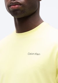 Κοντινό πλάνο ενός ατόμου που φοράει ένα ανοιχτό κίτρινο πουκάμισο Calvin Klein με στρογγυλή λαιμόκοψη, εμφανίζοντας το κάτω μέρος του προσώπου, τον λαιμό και το επάνω μέρος του στήθους.