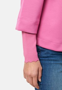 Sweat-shirt rose avec un poignet replié et un ourlet festonné sur la manche longue, porté avec un jean denim bleu. Tissu doux et texturé.