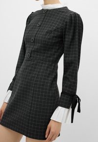 Robe ajustée gris foncé à carreaux, avec de gros boutons, des manches bouffantes, des poignets plissés blancs et des rubans noirs noués aux poignets.