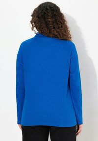 Camicia a maniche lunghe in un vivace tessuto blu con una texture liscia, caratterizzata da un design semplice e un orlo leggermente arrotondato sul retro.