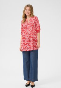 Rote Blumentunika mit V-Ausschnitt und dreiviertellangen Ärmeln; kombiniert mit weit geschnittenen Jeans und schwarzen High Heels. Heller Hintergrund.