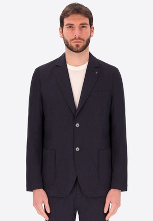 Uomo che indossa un blazer blu scuro con due bottoni, tasche applicate e una maglietta bianca girocollo su uno sfondo chiaro e uniforme.