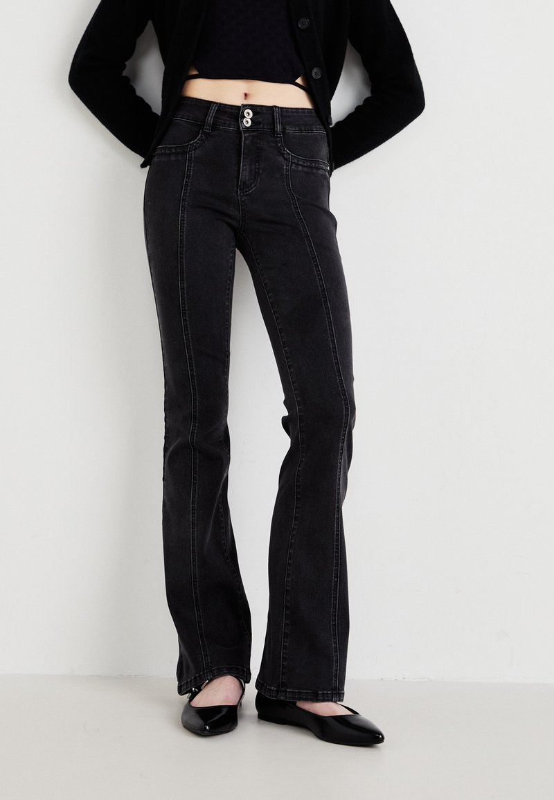 Calças jeans de brim preto com costuras brancas contrastantes, botões na frente e uma cintura baixa. Uma modelo as usa com sapatilhas pretas.