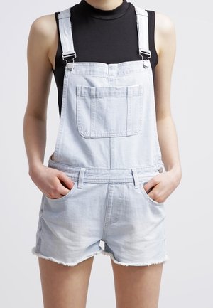 Person trägt hellblaue kurze Latzhosen aus Denim mit Vordertasche und schwarzes ärmelloses Oberteil, Hände in den Taschen, vor einfarbigem Hintergrund.