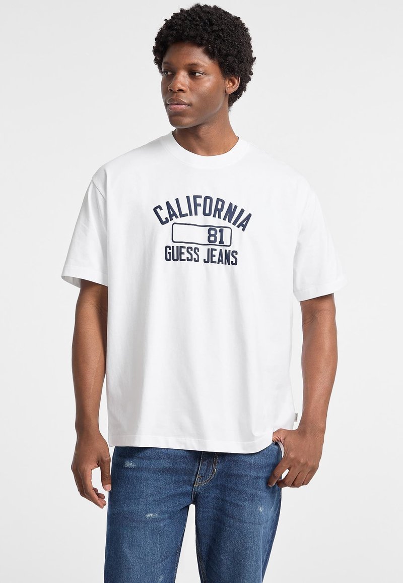 Valge puuvillane T-särk lühikeste varrukatega, millel on trükitud "CALIFORNIA 81" ja "GUESS JEANS" tumesinises värvuses. Kombineeritud siniste teksadega.