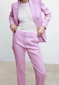 Frau in einem hellrosa Anzug, die mit den Händen ihre Hose zurechtrückt, kombiniert mit einem weißen gerippten ärmellosen Oberteil, stehend auf einem schlichten Boden.