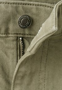 Nahaufnahme von khakifarbenem Denim-Stoff mit einem Metallknopf, der mit "MNG DENIM" beschriftet ist, und einem teilweise geschlossenen Reißverschluss am Bund eines Kleidungsstücks.