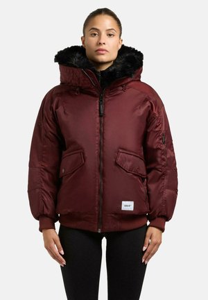 Giacca trapuntata color bordeaux con cappuccio foderato in pelliccia, zip frontale, polsini a coste e due tasche laterali. Presenta un'etichetta con logo sull'orlo.