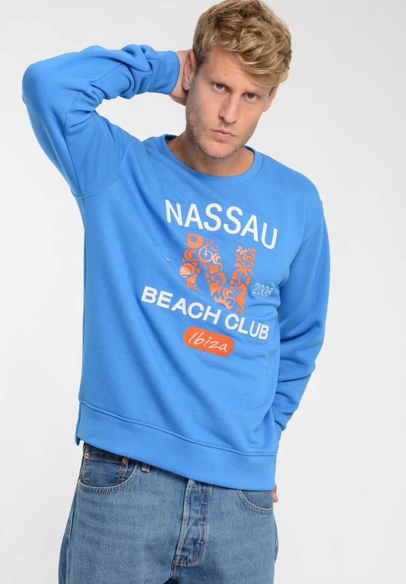 Jasnoniebieska bluza z rękawami i dołem wykończonym ściągaczem, z napisem "NASSAU BEACH CLUB" w białym i pomarańczowym kolorze oraz dekoracyjnymi wzorami.