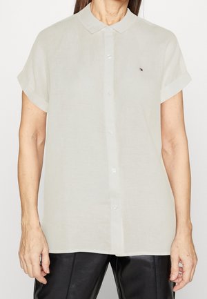 Femme portant une chemise crème à manches courtes boutonnée avec un petit logo sur la poitrine gauche, associée à un pantalon noir.