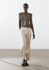 Crème-kleurige wijde broek met een gestructureerde bruine gebreide top en een zilveren ketting, gedragen met zwarte sandalen. Effen achtergrond.