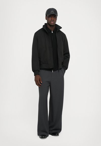 Jachetă bomber neagră cu guler înalt, combinată cu un top negru cu fermoar și pantaloni largi gri. O șapcă neagră finalizează look-ul.