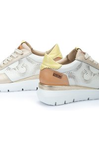 Sneakers blancos y beige con diseño perforado, detalles en dorado, tacón beige y lengüeta trasera texturizada amarilla sobre una suela blanca.