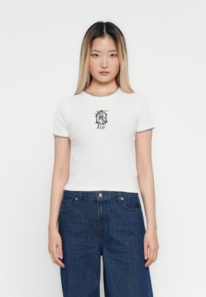 Giovane donna con capelli lisci ombre, che indossa una maglietta bianca aderente con una grafica nera e jeans a gamba larga blu scuro, in piedi davanti a uno sfondo bianco.