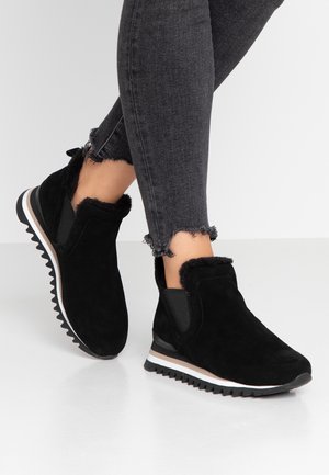 Pieds chaussés de bottines en daim noir avec doublure en fourrure et semelles à rainures, assorties à un jean skinny gris foncé à ourlet effiloché.
