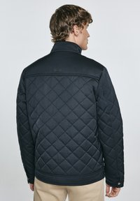 Giacca nera trapuntata con colletto alto, caratterizzata da un motivo a diamante testurizzato e un discreto patch con logo sulla schiena.