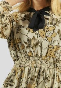 Frau, die ein beiges und schwarzes Blumenkleid mit einer schwarzen Schleife am Hals und elastischem Bund trägt.