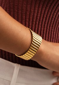 Pulsera de brazalete texturizado ondulado dorado llevada en una muñeca, sobre una blusa de tono rojo oscuro con acanaladuras y pantalones de color claro.