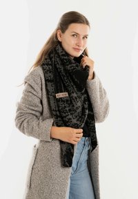 Zwarte en grijze gebreide sjaal met franjes, gedragen over een grijze gestructureerde jas. Bevat een klein bruin label met "Knit Factory."