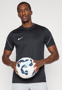 Μαύρο αθλητικό μπλουζάκι Nike με λευκές λεπτομέρειες, κρατημένο με μια λευκή και μπλε κα patterned μπάλα ποδοσφαίρου, φορεμένο με ανοιχτό γκρι σορτς, κοντά μανίκια.