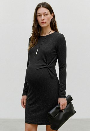 Femme enceinte vêtue d'une robe noire ajustée à manches longues, tenant une grande pochette noire, avec de longs cheveux bruns et des bijoux minimalistes.