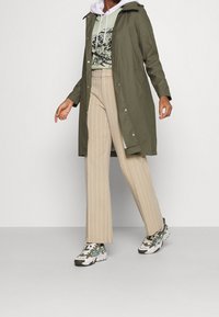 Oliven grøn lang frakke med hætte, beige stribede bukser og mønstrede sneakers, med et grafisk print på den skjorte, der er under.