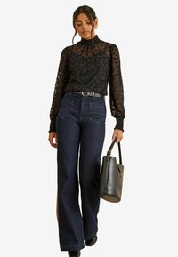 Blusa nera a pois con texture trasparente, colletto alto e maniche a sbuffo abbinata a jeans larghi scuri e una borsa nera.