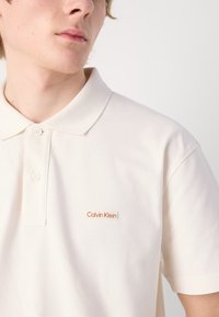 Krēmveida polo krekls ar mīkstu tekstūru, trīs pogu plāksne un kontrastējošs "Calvin Klein" logo oranžā krāsā kreisajā krūtī.