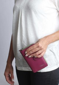 Portafoglio in pelle bordeaux con dettagli verdi, dotato di chiusura a zip e bottone a scatto anteriore, tenuto in una mano su uno sfondo chiaro.