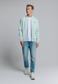 Lichtblauwe bomberjack, met rits, ribbelmanchetten en onderrand, gedragen over een wit t-shirt, in combinatie met verwasde blauwe jeans en witte sneakers.