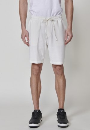 Man in witte gestructureerde drawstring shorts, een wit T-shirt en zwarte sneakers die tegen een effen achtergrond staat.