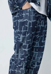 Pantalons en denim bleu avec un motif texturé géométrique dans une teinte plus sombre. Le tissu est doux, avec une coupe décontractée et des poches latérales.