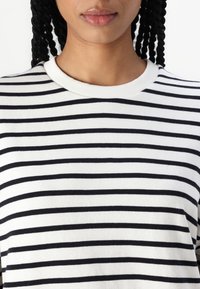 Camiseta blanca con rayas negras horizontales, cuello redondo acanalado, tela suave y ajuste relajado. Presenta un patrón uniforme de rayas en toda la prenda.