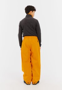 Fleece coltrui in donkergrijs, gecombineerd met loszittende oranje broeken met een elastische tailleband en zwarte laarzen.