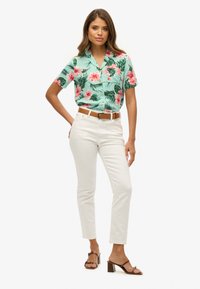 Camicia a maniche corte azzurro chiaro con motivi floreali rosa e foglie verdi, abbinata a pantaloni bianchi aderenti e sandali con tacco marroni.