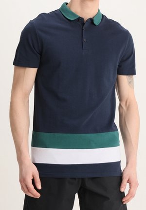 Homme portant un polo bleu marine avec un col vert foncé et des rayures horizontales vertes et blanches près de l'ourlet, associé à un pantalon noir.
