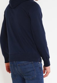 Sweat à zip bleu marine, en mélange de coton, avec une capuche et des poignets côtelés. Porté avec un jean bleu ajusté, montrant un minimum de détails.