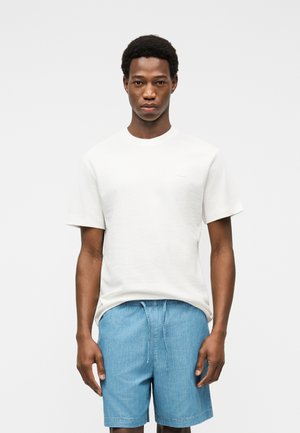 Jeune homme portant un t-shirt blanc uni à manches courtes et un short bleu clair à cordon, debout devant un fond uni.