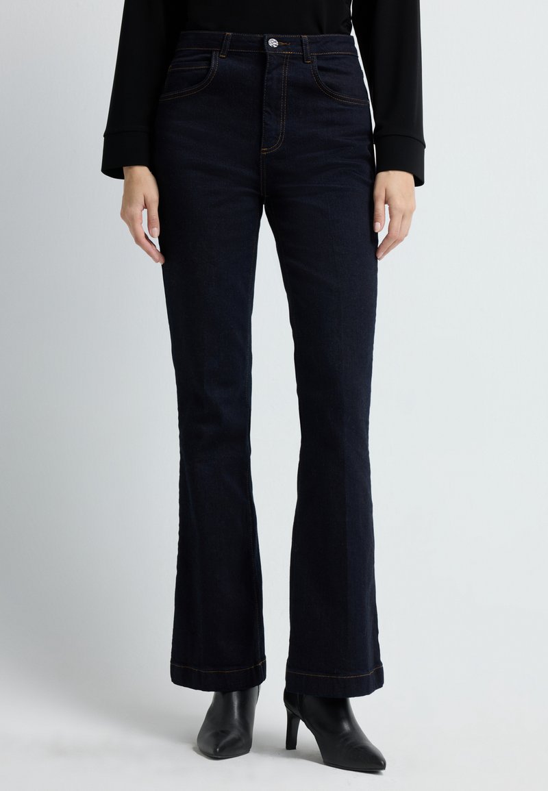 Tummat denimfarkut ovat leveälahkeiset, korkealla vyötäröllä, viidellä taskulla ja niissä on kontrastina oranssia tikkausta. Kangas näyttää sileältä ja siinä on hieman kiiltoa.