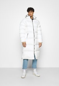 Giovane uomo che indossa un lungo cappotto bianco in piuma con cappuccio, jeans azzurri, sneakers bianche, in piedi contro uno sfondo bianco uniforme.