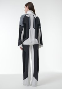 Vrouw met haar rug naar de camera, gekleed in een zwart, wit en grijs hoge hals top en wijde broek met geometrische kleurblokken op een witte achtergrond.