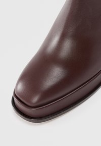 Bottine en cuir marron avec une finition lisse et brillante, bout rond et un léger talon. Présente un discret détail de couture le long du bord.