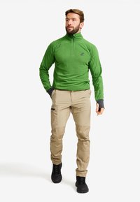Green fleece pullover with half-zip, paired with light tan pants featuring zip pockets and contrasting black cuffs. Black footwear.